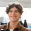 Smiling young man with curly hair wearing a brown jacket over a white shirt, sitting indoors in a softly lit workspace with blurred background.