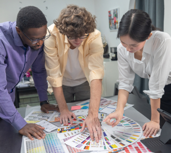 Three diverse professionals collaborating around a desk, reviewing color swatches, branding charts, and printed design samples in a modern office, focused on visual identity planning and creative strategy.