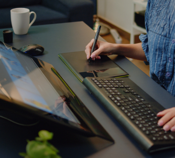 A graphic designer uses a digital pen and tablet at a modern workstation, working alongside a desktop computer and keyboard, illustrating digital illustration, graphic design, and creative content creation in a professional workspace.