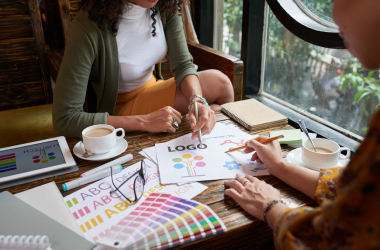Two designers collaborate at a café table, reviewing logo concepts and brand identity materials with color palettes, typography samples, and sketches, while working on visual branding strategy in a creative workspace with coffee cups nearby.