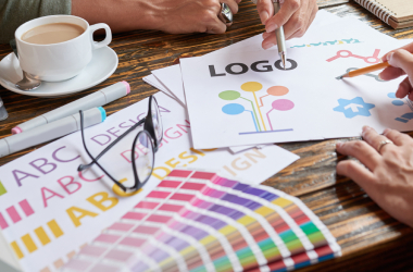Designers work on logo creation and brand identity planning, reviewing logo sketches, color palette swatches, and typography samples spread across a wooden table with coffee cups, representing creative branding and visual design collaboration.
