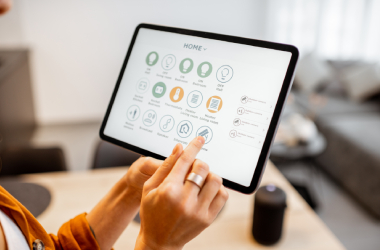 Two UX/UI designers collaborate in a modern office, reviewing mobile app wireframes on a tablet and sketching interface layouts on paper, with a computer screen displaying UX/UI design mockups in the background, highlighting user experience and interface design planning.